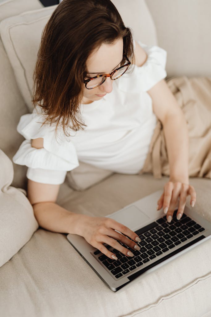 A woman in glasses types on a laptop while lounging on a couch, exemplifying modern lifestyle and remote work.
